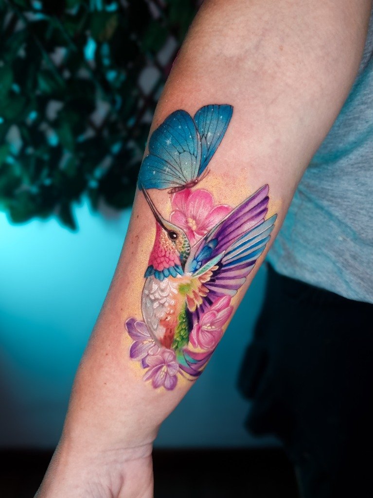 Tatuaje a color de colibrí y mariposa azul en el antebrazo